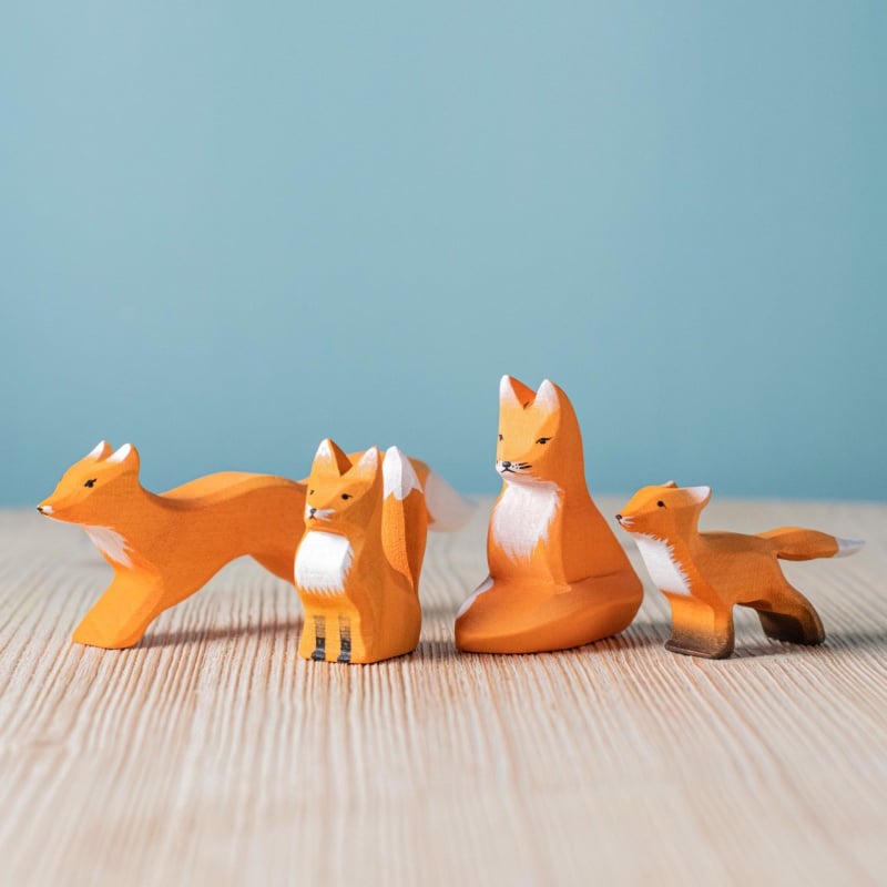 Four orange wooden fox figures in various sizes and poses are displayed on a light wooden surface with a blue background—perfect as the Bumbu Handmade Wooden Fox Family Set of charming handmade toys.