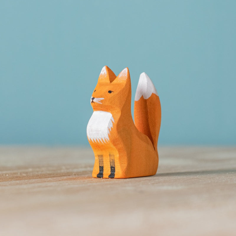 A small fox figurine from the Bumbu Handmade Wooden Fox Family Set, featuring orange and white painted details, sits on a light wooden surface against a pale blue background. Handmade in Romania.
