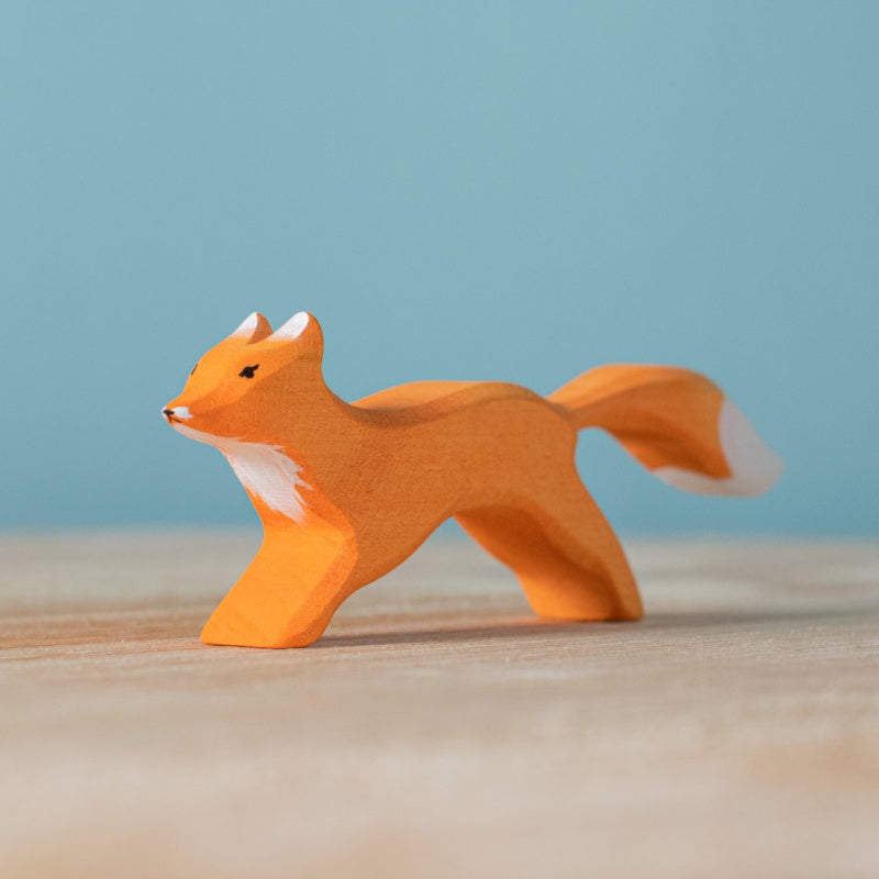 An orange-and-white fox from the Bumbu Handmade Wooden Fox Family Set, crafted in Romania, stands on a light wood surface with a blue background.