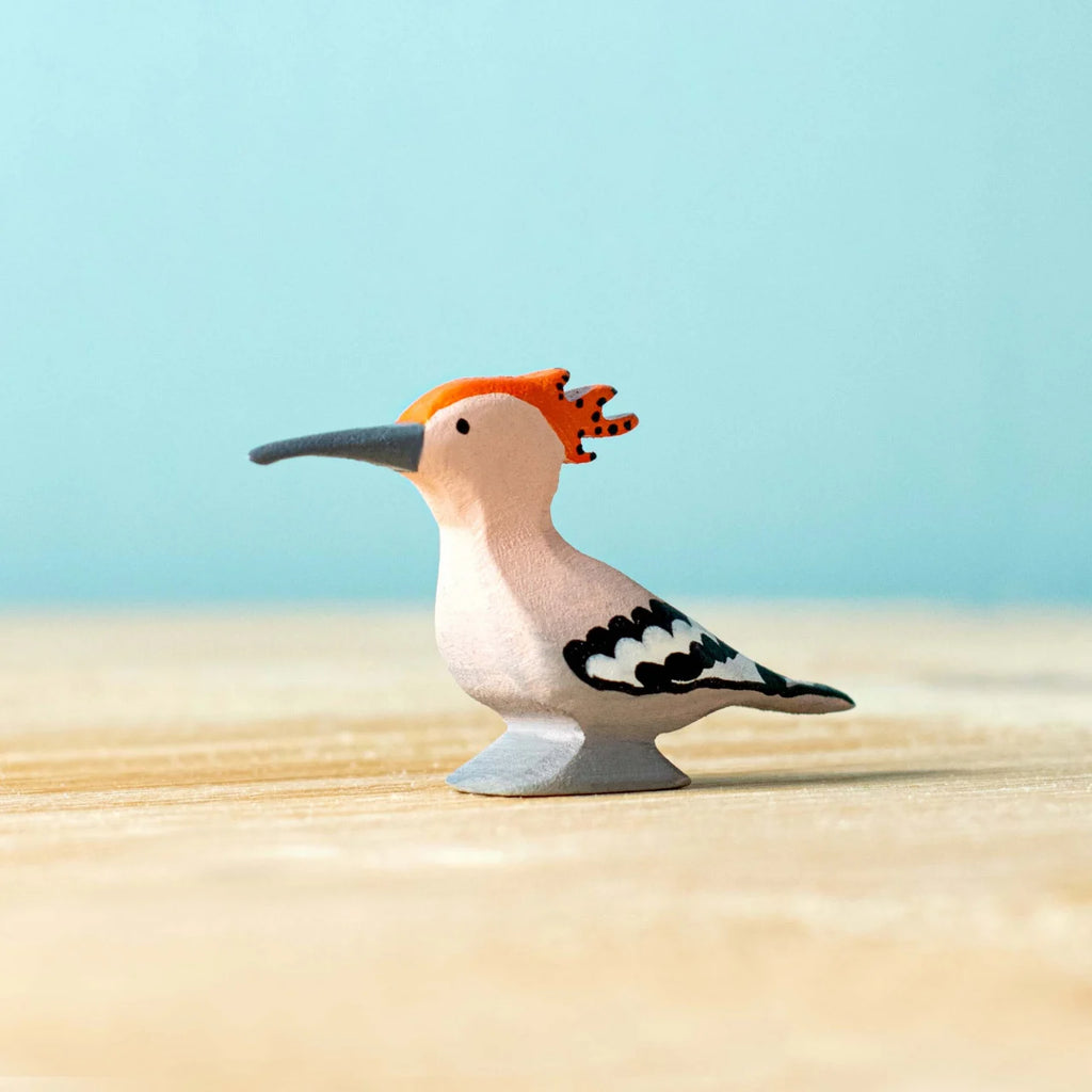 The Bumbu Handmade Wooden Hoopoe Bird, an eco-friendly handcrafted toy with an orange crest, black beak, and black-and-white wings, stands on a light wooden surface against a pale blue background.