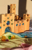 A Bauspiel Large Fairytale Castle, 16-Piece Set made of wood with crenellated towers and inlaid star and circular gems sits on colorful fabric scattered with translucent cubes designed for imaginative play.