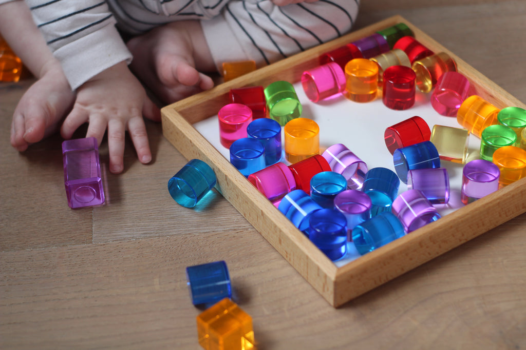 A child sits beside a wooden tray filled with colorful, translucent Bauspiel Lucent Cylinders, 64-Piece Set.