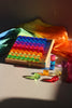 A wooden tray holding an array of sixty-four translucent, rainbow-colored Bauspiel Lucent Cylinders, with several extra pieces from the set resting on the surface nearby.