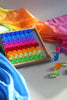 A wooden tray filled with rows of translucent, rainbow-colored Bauspiel Lucent Cylinders, 64-Piece Set, with a stack and a few loose pieces beside it, set against colorful fabric.