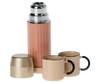 A pink thermos with a shiny silver cap from the Maileg Camping Set stands like a beacon. Beside it, two beige cups with black handles rest quietly. The cap, turned upside down in front of the thermos, resembles another cup ready for adventure.