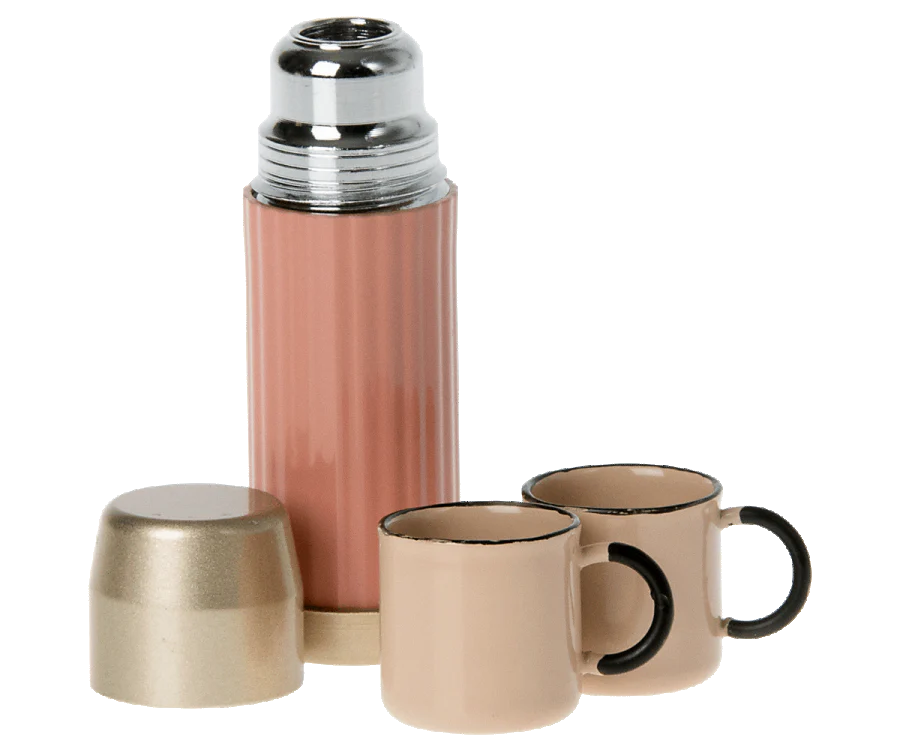A pink thermos with a shiny silver cap from the Maileg Camping Set stands like a beacon. Beside it, two beige cups with black handles rest quietly. The cap, turned upside down in front of the thermos, resembles another cup ready for adventure.