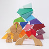 A colorful arrangement of wooden toys forming a creative structure. The vibrant Grimm's Rainbow Lion Building Set includes pieces in various colors like green, blue, purple, red, orange, and yellow. In the foreground are natural wood figures resembling abstract human forms and geometric shapes.