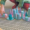 A child wearing a green shirt and red polka dot pants plays with the Pastel Semi Circle Set on a patterned rug, reminiscent of modern architecture. The shapes include cylinders, cones, and discs in pastel shades of blue, pink, and purple, arranged in a symmetrical pattern.