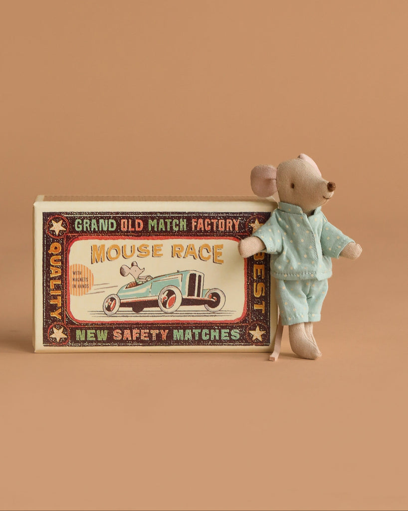 A Maileg Mouse Family Collection figure in blue pajamas rests against a vintage-style "Mouse Race" matchbox with a race car design, all set on a plain beige background.