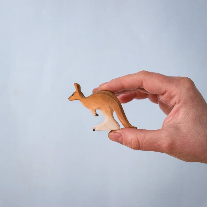 A hand holds a small Bumbu Wooden Kangaroo Family Set figurine—crafted from sustainably sourced wood—against a plain, light blue background.
