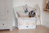 A white wicker bench with a cushion, decorated with a swan plush, llama doll, and the Hot Air Balloon - Light Brown / Navy Blue, sits in the corner of a children's room with light wood floors and neutral furniture.