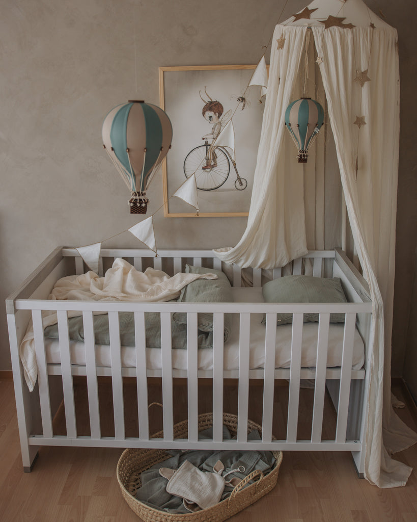 A white baby crib with green cushions and blanket features a Hot Air Balloon – Pastel Green mobile, a canopy with gold stars, handmade children’s decor like a wooden balloon ornament, framed illustration, and a wicker basket under the crib.