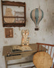 A child's desk with an open storybook, cat-shaped lamp, and wooden chair. Above are a pastel green Hot Air Balloon decoration, an alphabet plaque, shelf signs, all set against floral wallpaper.