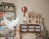 A cozy children's room featuring shelves of books, a wooden dollhouse, storage bins, a hanging plush swan, the Hot Air Balloon - Burgundy decor, a tiger-shaped cutout, and a doll with a pointed hat sitting on a shelf.