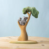 The Bumbu Handmade Wooden Koala Bears with Eucalyptus Tree Set features an adult and baby koala on green-leaved branches, displayed on a light wooden surface against a blue background.