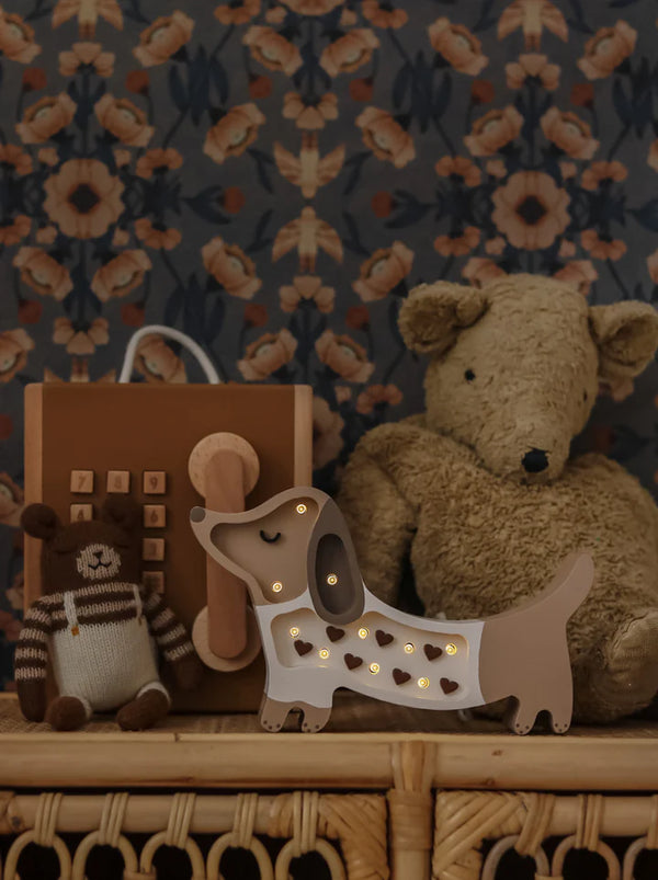 A cozy nursery scene featuring a plush teddy bear, a crocheted dog toy, a brown toy telephone, and the Handmade Wooden Mini Lamp - Puppy on a wicker shelf—perfect decor set against dark floral wallpaper.
