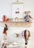 A wooden shelf displays a red teapot set, white cone trees, a cherry print, and a stuffed mouse. Below hang a toy camera, scarf, small wooden airplane, and Hot Air Balloon - Light Brown / Dark Brown for children's room decor.