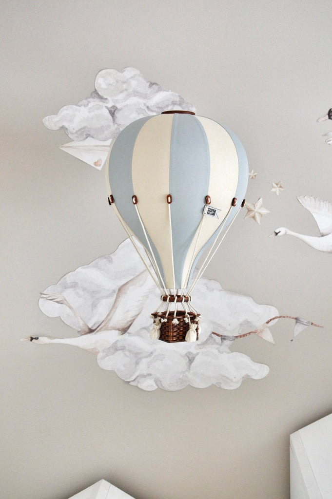 The Hot Air Balloon - Beige / Mint / Grey features soft stripes and a wicker basket, floating against a mural of clouds, paper planes, and swans. This handmade children’s room decoration adds charm to any dreamy space.