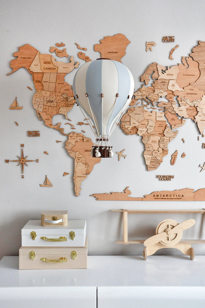 A decorative wooden world map hangs on the wall behind the Hot Air Balloon - Beige / Mint / Grey model. Below, a white dresser displays stacked suitcases and a wooden toy airplane, creating a charming children's room decor.