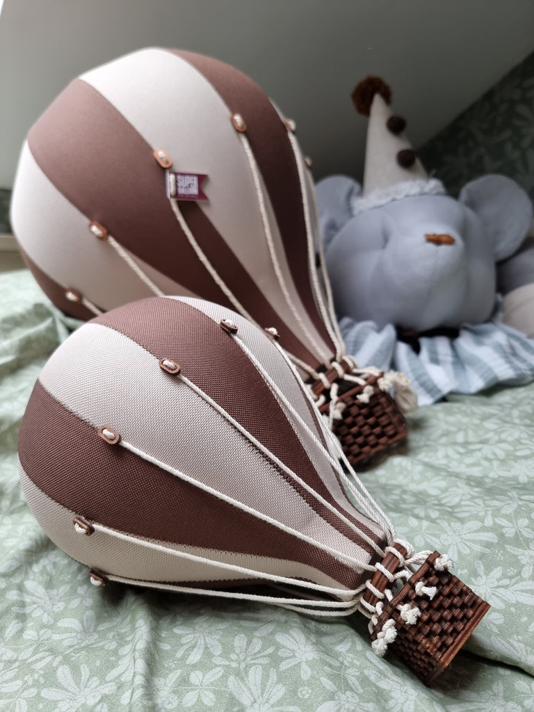 Two handmade Hot Air Balloon decorations in Light Brown and Dark Brown fabric and wicker rest on a green patterned surface. A plush elephant in a clown hat sits behind them—ideal accents for a charming children's room.