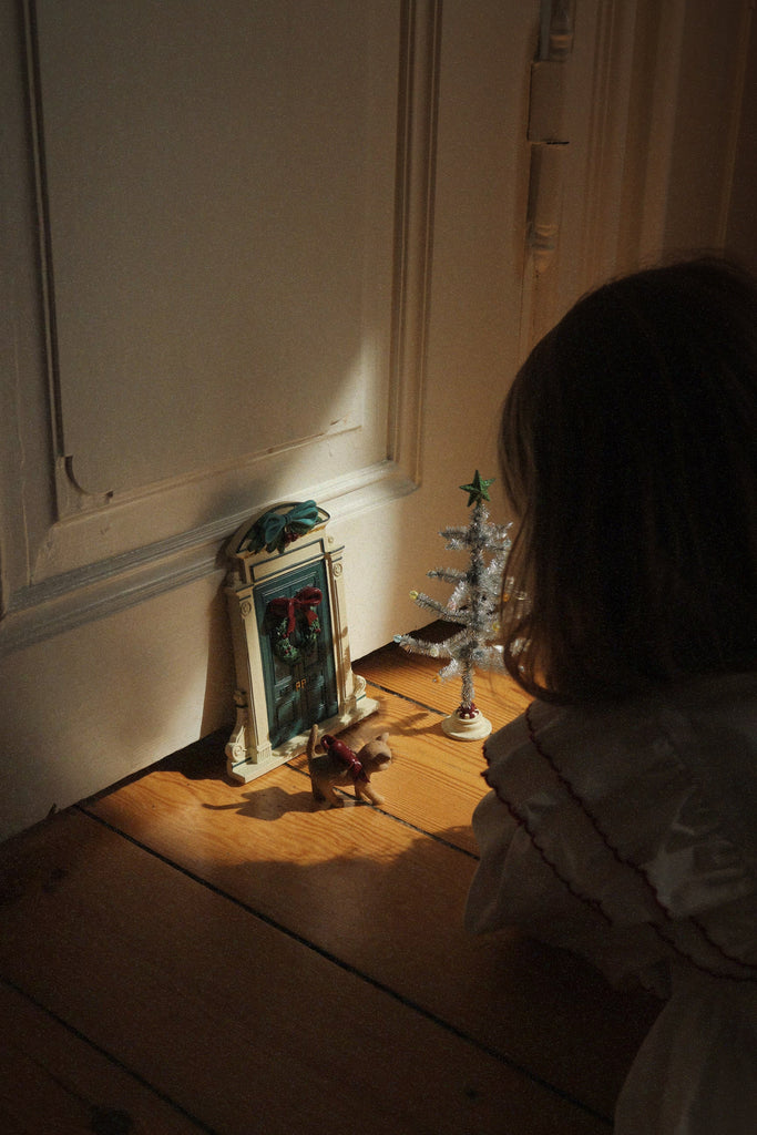 A child looks at a Konges Sloejd Christmas Door, a small decorated tree, and a toy cat placed on a wooden floor.