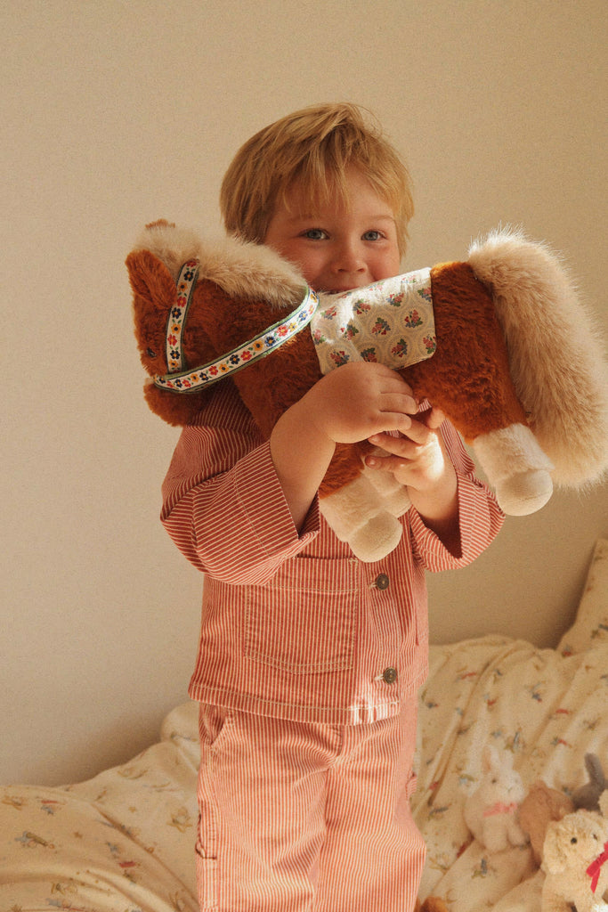 A young child in striped pajamas hugging a Konges Sloejd Fiona Horse with a floral bridle and saddle.