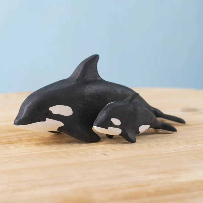 The Bumbu Handmade Wooden Orca Set features two black-and-white orca figurines, one large and one small, displayed on a light wooden surface against a pale blue background—ideal sustainable toys for any Montessori playset.