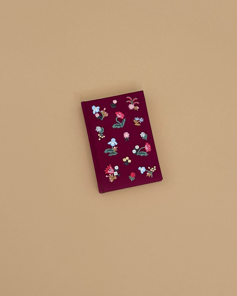 The Gracie Mini Embroidered Journal, a red hardcover with floral and berry embroidery, rests on a plain light brown surface and features blank unlined pages for creative expression.