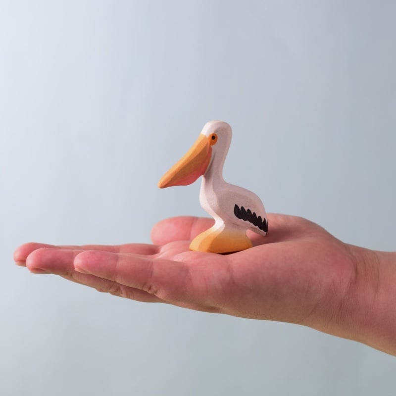 A hand holds a Bumbu Handmade Wooden Pelicans Family Set piece—a small wooden pelican with an orange beak and yellow base, designed for Montessori play—set against a plain light blue background.