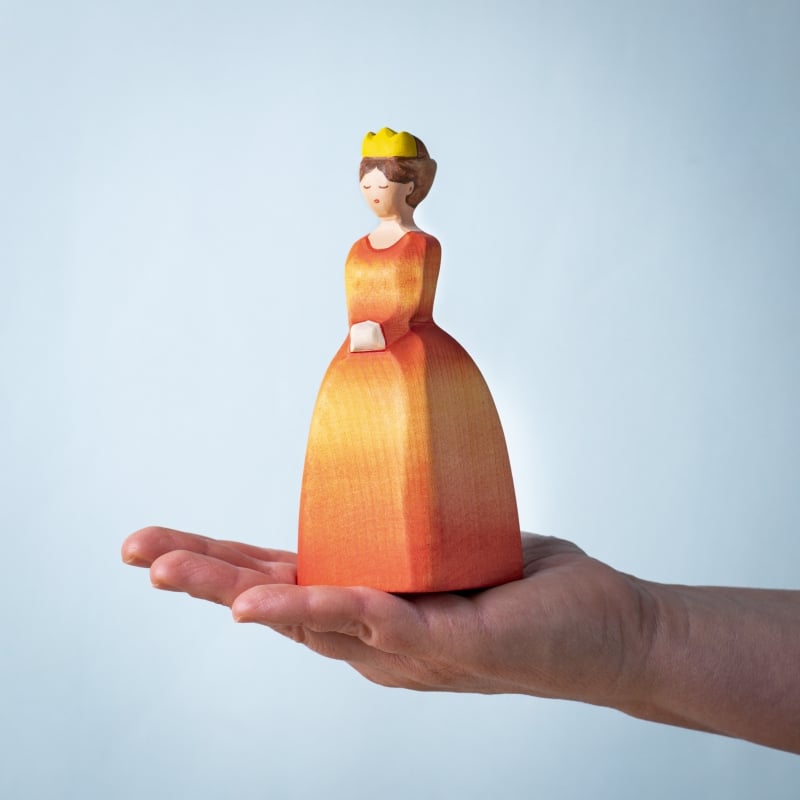 The Bumbu Handmade Wooden Princess, featuring a woman in a yellow-orange dress and crown, is balanced on an open palm against a plain light blue background.