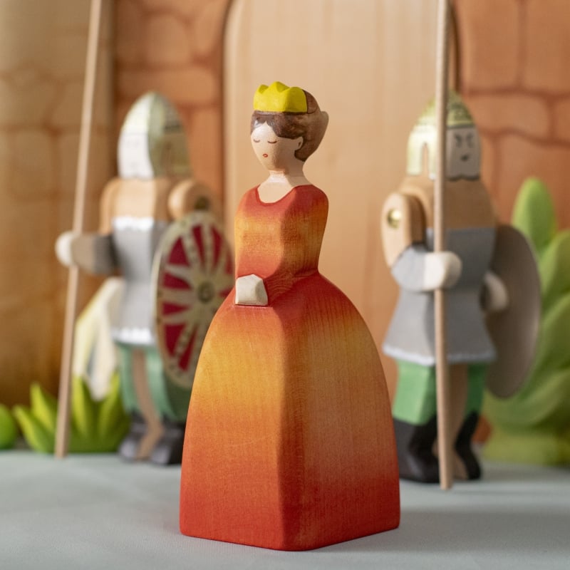The Bumbu Handmade Wooden Princess—sporting an orange dress and yellow crown—stands before two wooden knights with spears and shields, ideal for Waldorf-inspired play and eco-friendly toy collections.