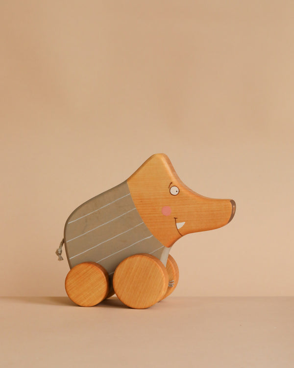 The Pull and Push Toy Little Wild Boar is a handcrafted wooden toy with a grey painted body, round wooden wheels, rosy cheeks, and made from natural materials. Set against a plain beige background.