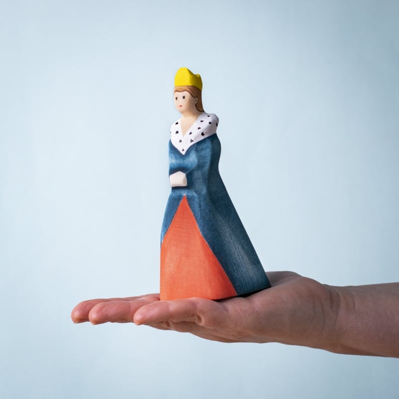 A hand holds the Bumbu Handmade Wooden Queen, featuring a yellow crown, blue and red dress, and a white cape with black spots, set against a light blue background.