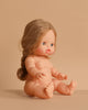 The Minikane Baby Doll (13") - Eleanor, featuring long light brown braided hair and blue eyes, sits unclothed with outstretched arms and bent legs on a beige surface against a beige background. Clothing sold separately.