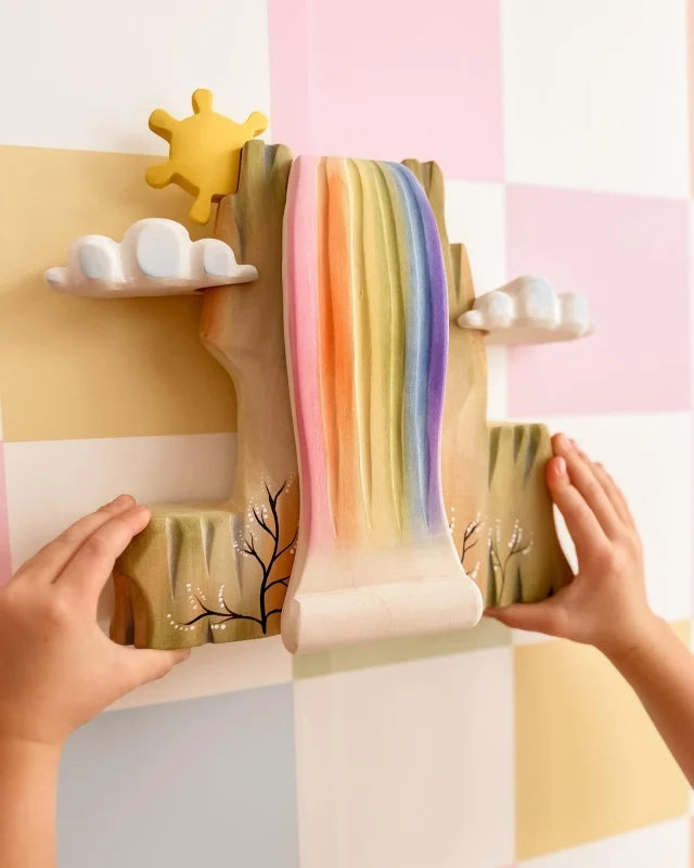 A child’s hands touch the Bumbu Wooden Rainbow Cliff—a colorful wall decor featuring a rainbow waterfall, clouds, and sun on a pastel checkered backdrop with painted tree branches—perfect for imaginative play. Ships in one week.