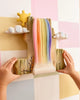 A child’s hands touch the Bumbu Wooden Rainbow Cliff—a colorful wall decor featuring a rainbow waterfall, clouds, and sun on a pastel checkered backdrop with painted tree branches—perfect for imaginative play. Ships in one week.