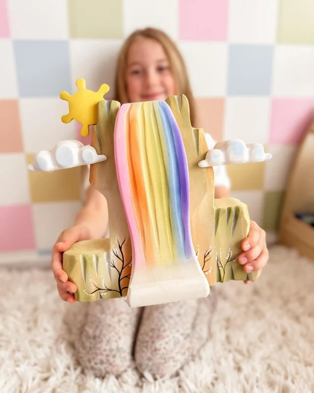 A smiling girl sits on a fluffy rug, holding the Bumbu Wooden Rainbow Cliff—made from sustainably sourced wood with clouds and a sun—perfect for imaginative play, against a pastel checkered wall. (Ships in one week.).