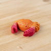The Bumbu Handmade Wooden Red Crab, a small handcrafted figurine with red claws and white spots, sits on a light wooden surface.