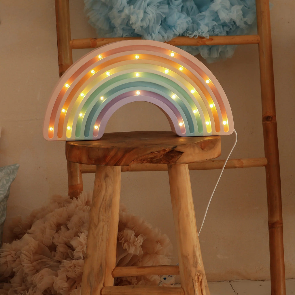 The Little Lights Rainbow Lamp, a handmade children's night light with glowing pastel colors, sits on a wooden stool in front of a ladder adorned with fluffy blue and beige fabric.