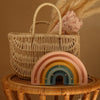 A woven basket with a floral cloth rests on a rattan table. In front, the Handmade Wooden Mini Lamp - Rainbow, a pastel-colored night light with gold accents, softly glows against the warm beige background.