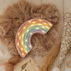 The Little Lights Rainbow Lamp, a pastel-colored children's night light with small LEDs, sits on beige ruffled fabric and is surrounded by a woven rattle and floral-patterned decor.