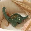 The Handmade Wooden Mini Lamp - Dinosaur features a green dinosaur design with small yellow bulbs, placed in a beige basket next to dried pampas grass and a brown pillow—an adorable touch for your nursery decor.