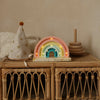 A Handmade Wooden Mini Lamp - Rainbow sits on a wicker table beside a star-patterned party hat, wooden stacking rings, and the book "Who am I?", its gentle glow softly illuminating the neutral wall in this charming children's scene.