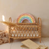 The Little Lights Rainbow Lamp, a handmade pastel rainbow-shaped children's night light, sits in a small wicker crib with a beige cushion. Dried pampas grass rests on the crib amid scattered cards, all against a cream-colored wall.