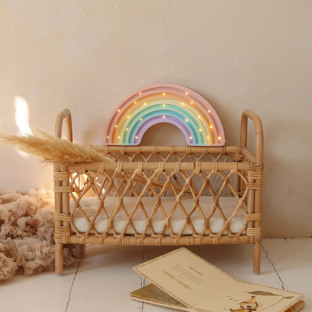 The Little Lights Rainbow Lamp, a handmade pastel rainbow-shaped children's night light, sits in a small wicker crib with a beige cushion. Dried pampas grass rests on the crib amid scattered cards, all against a cream-colored wall.