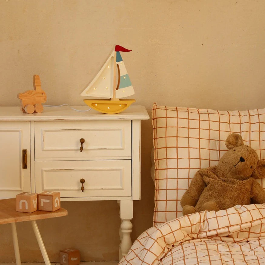 A cozy children's bedroom features a white nightstand with a Handmade Wooden Lamp - Mini Sailboat, Mustard, and bunny figurine. The orange grid bedding sits beside alphabet blocks and a teddy bear—ideal for ocean-themed nursery decor.