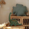 A Handmade Wooden Mini Lamp - Dinosaur, with small glowing accents, sits on a wicker table near a rattan shelf. Above, a green leaf-shaped cushion adds cozy charm—ideal nursery decor for a natural-themed space.