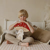 A young blond child sits cross-legged on a bed, smiling and holding a large wooden mushroom toy. Nearby, the Handmade Wooden Lamp - Mushroom House made of natural pinewood adds charm to the cozy, neutral-toned room.