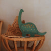The Handmade Wooden Mini Lamp - Dinosaur adds whimsical charm to nursery decor, standing beside a pinewood mountain puzzle on a round wicker table against a beige wall.
