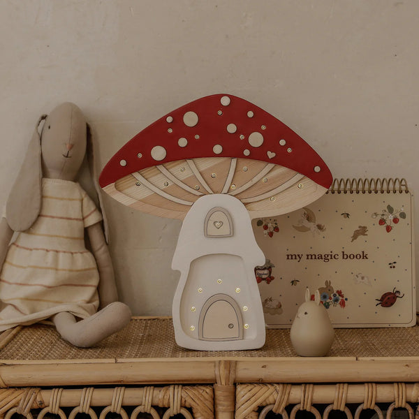A plush rabbit doll, the Handmade Wooden Lamp - Mushroom House, a beige "my magic book," and a small figurine sit on a rattan shelf against a neutral wall—an ideal children’s night light display.